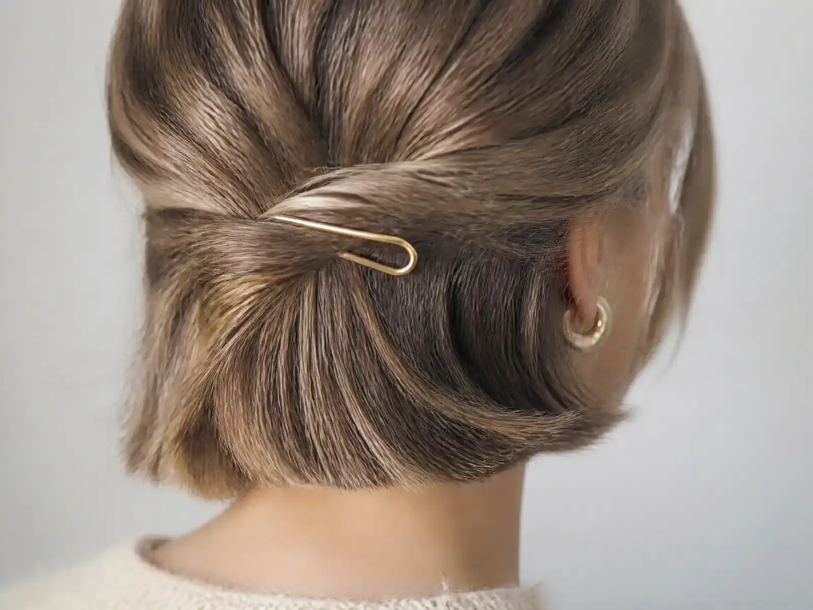 A small gold U-shaped French hair pin holding a compact low twist in chin-length dark blonde hair, the loose ends of the bob falling free below it