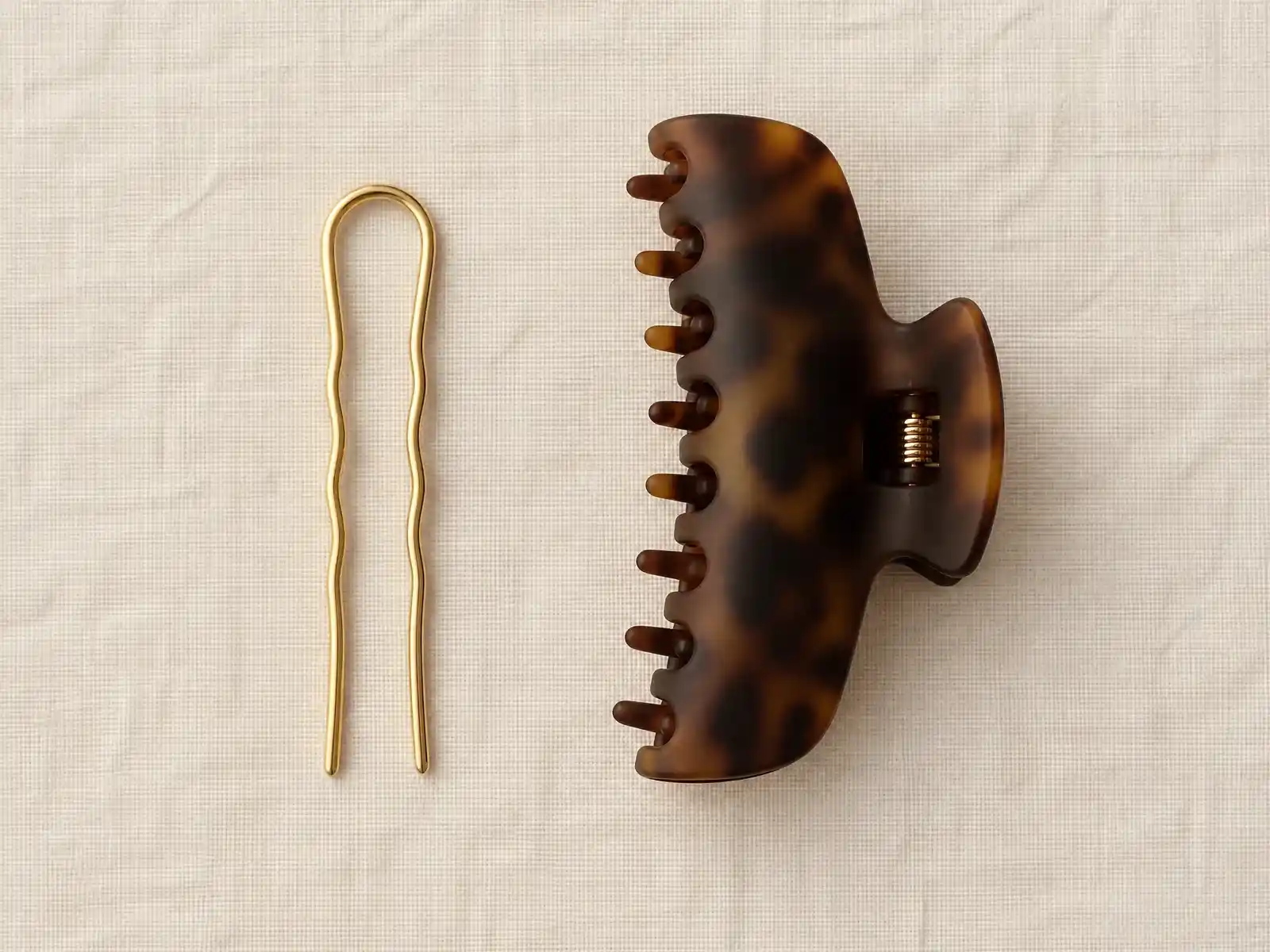 A slim gold U-shaped French hair pin and a large matte tortoiseshell claw clip laid side by side on warm neutral linen for comparison