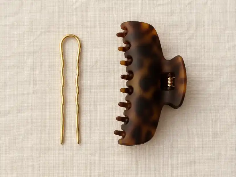 A slim gold U-shaped French hair pin and a large matte tortoiseshell claw clip laid side by side on warm neutral linen for comparison