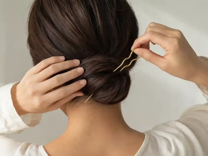 Two hands setting a wavy gold French hair pin into a smooth low chignon of dark brown hair, the pin caught partway through its rotation into the base of the bun