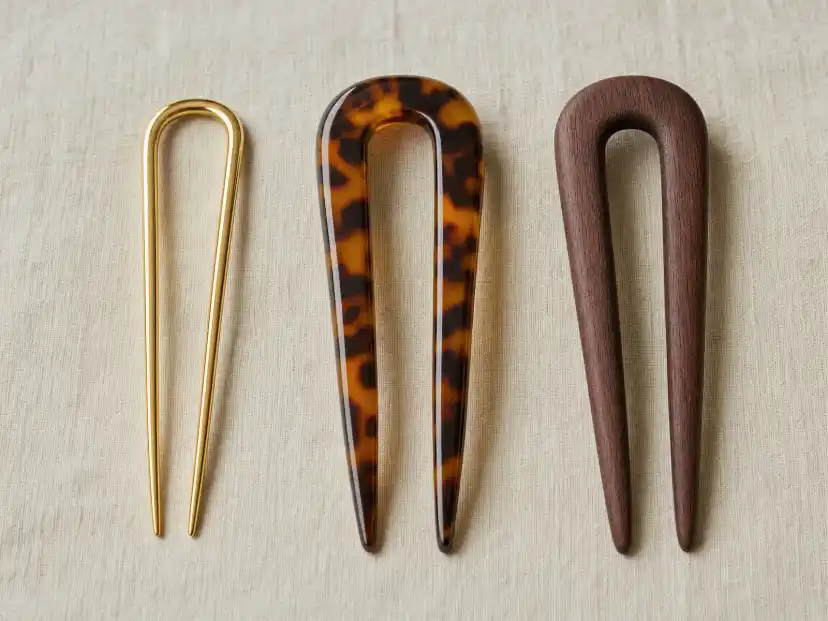 A polished gold metal French hair pin, a glossy tortoiseshell acetate pin and a matte rosewood pin laid side by side on neutral linen, showing the difference in thickness and surface finish