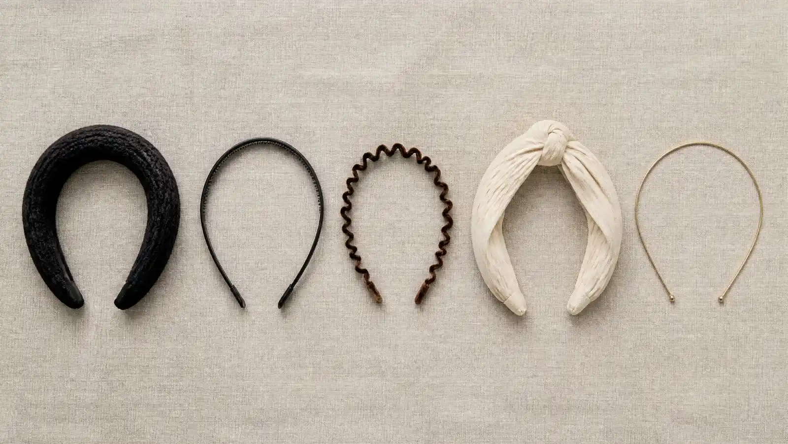 An overhead row of different headband types on linen: a wide padded knit band, a toothed plastic hoop, a slim velvet band, a knotted wrap and a thin wire band